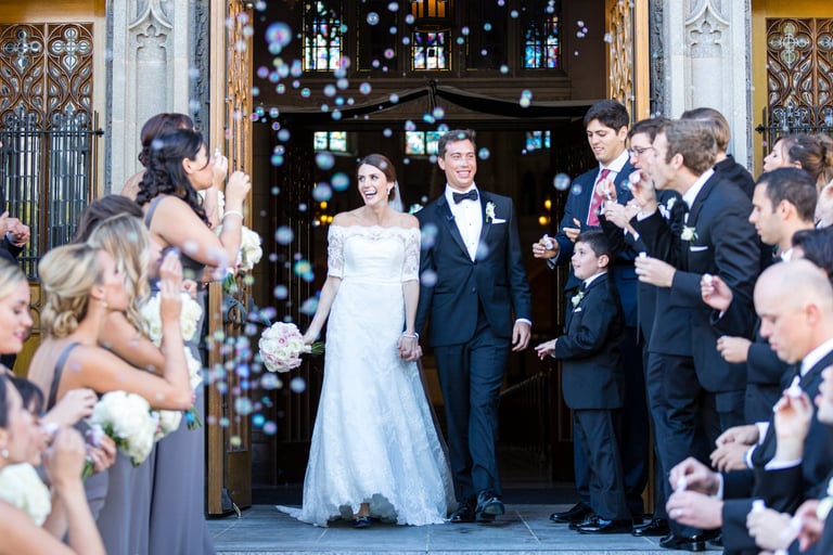 Bride and groom exit ornate church doors while guests throw confetti and bubble wands during wedding ceremony